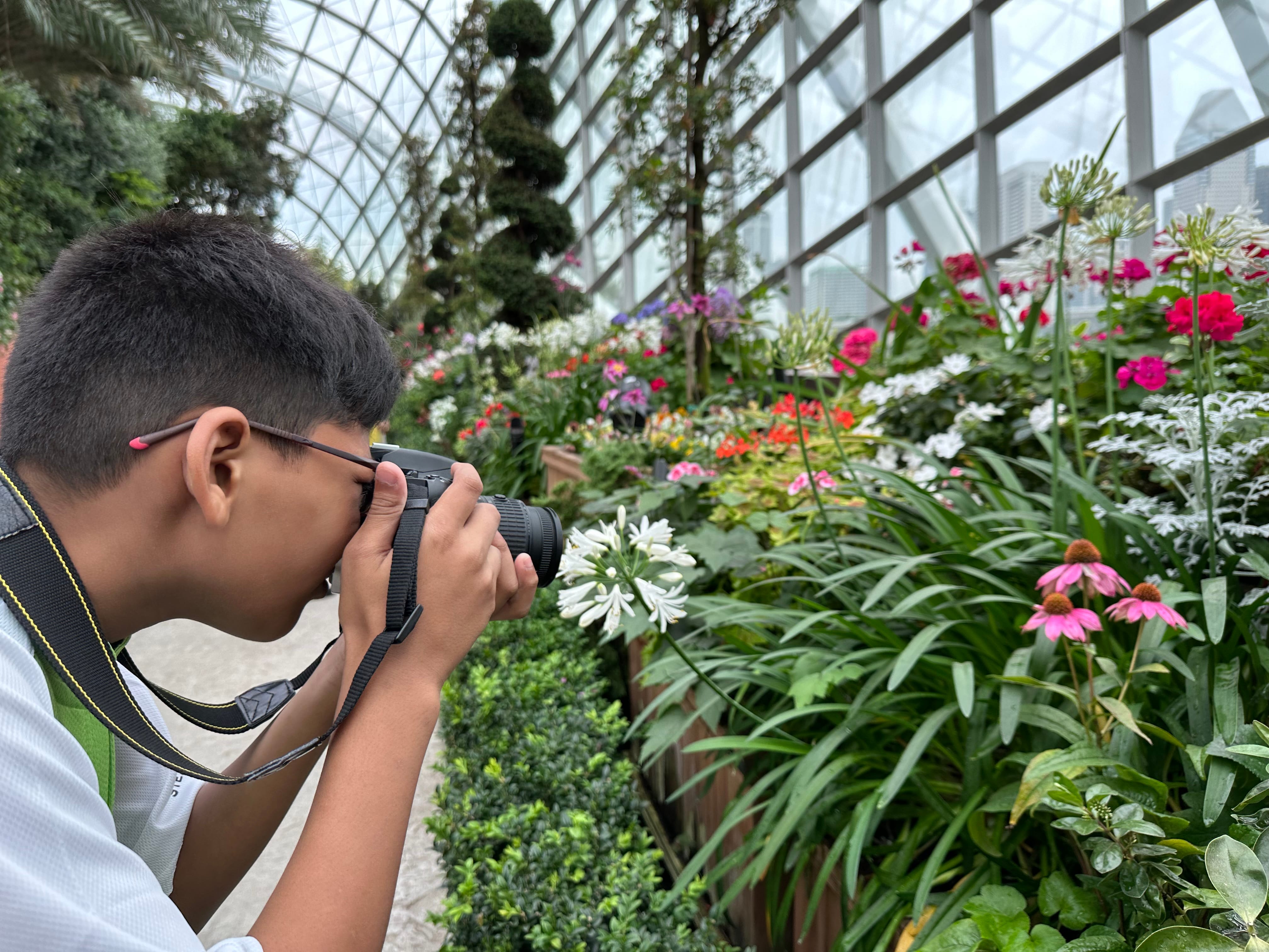 A boy with glasses photographs colorful flowers inside a large glass greenhouse dome.